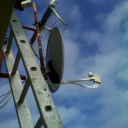 Antena satelitarna zamontowana na maszcie, widoczna z dołu, drabina oparta o maszt, niebieskie niebo w tle.