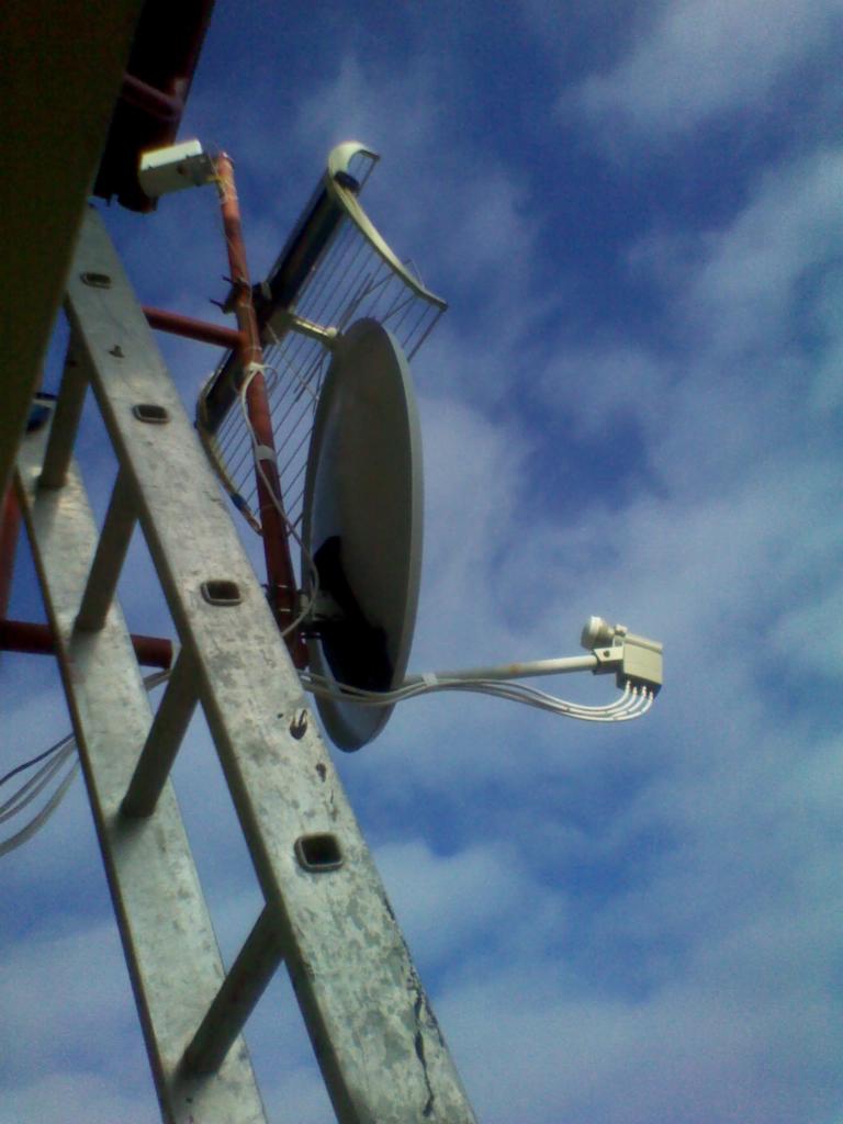 Antena satelitarna zamontowana na maszcie, widoczna z dołu, drabina oparta o maszt, niebieskie niebo w tle.
