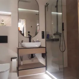 Nowoczesna łazienka z owalnym lustrem w czarnej ramie, umywalką nablatową na drewnianej szafce, przeszkloną kabiną prysznicową z czarną armaturą i oświetleniem LED pod szafką i wzdłuż kabiny.