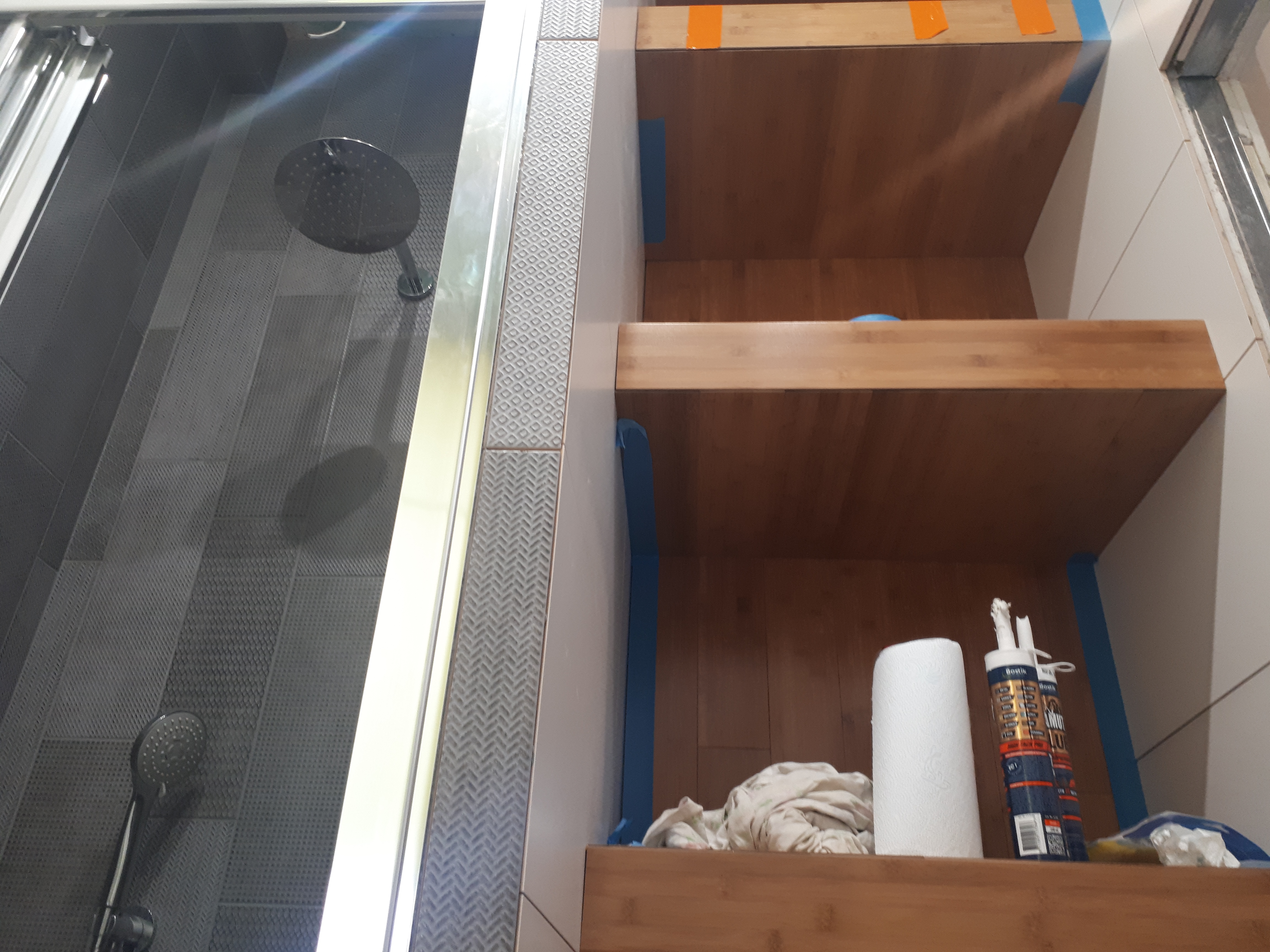 Półki z drewna bambusowego w trakcie montażu w łazience, zabezpieczone niebieską taśmą malarską, widoczne tuby z silikonem, umywalka i kabina prysznicowa z szarą mozaiką.