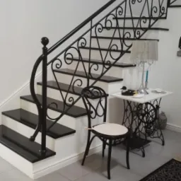 Eleganckie schody z czarnymi stopniami i ozdobną, kutą, czarną balustradą z motywem spiralnym, obok stolik z lampką i krzesłem w stylu retro.