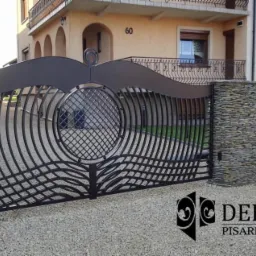 Nowoczesna brama wjazdowa z metalu o falistym wzorze, osadzona na kamiennych słupkach, na tle domu z numerem 60, żwirowe podłoże, widoczne logo firmy DEKOR-MET.