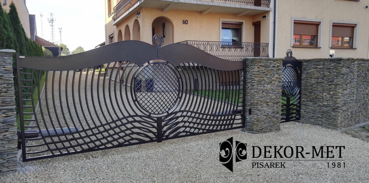 Nowoczesna brama wjazdowa z metalu o falistym wzorze, osadzona na kamiennych słupkach, na tle domu z numerem 60, żwirowe podłoże, widoczne logo firmy DEKOR-MET.