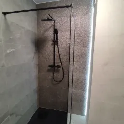 Nowoczesna kabina prysznicowa typu walk-in z czarną armaturą i brodzikiem, wykończona płytkami w odcieniach szarości i bieli, z subtelnym oświetleniem LED akcentującym strukturę ściany.