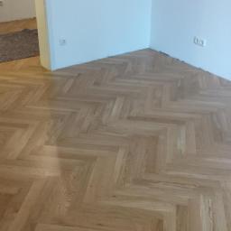 Renobud - Ułożony parkiet w jodełkę w jasnym odcieniu drewna, widoczne gniazdka elektryczne na białej ścianie i fragment innego pomieszczenia z wykładziną w tle.