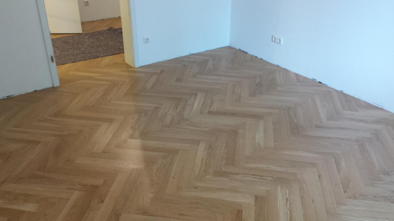 Ułożony parkiet w jodełkę w jasnym odcieniu drewna, widoczne gniazdka elektryczne na białej ścianie i fragment innego pomieszczenia z wykładziną w tle.