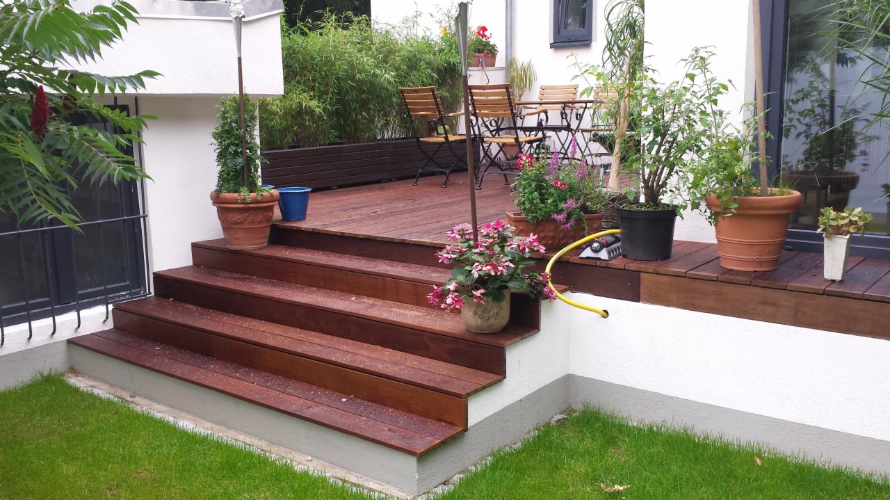 Drewniany taras z schodami, otoczony zielenią, z meblami ogrodowymi i doniczkami z kwiatami, widok na schludny trawnik.
