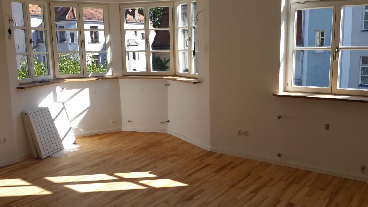 Jasne, puste wnętrze po remoncie z drewnianą podłogą, dużymi oknami i białymi ścianami, na których widać ślady po wcześniejszych mocowaniach.