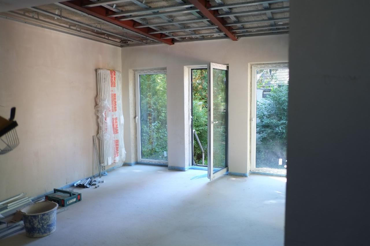 Pokój w trakcie prac wykończeniowych: surowa ściana z grzejnikiem w folii, odsłonięta konstrukcja sufitu, okna balkonowe z widokiem na zieleń, wiadro i narzędzia na podłodze.