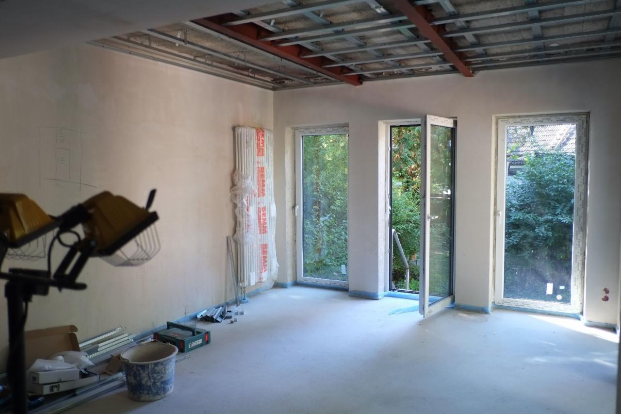 Wnętrze w trakcie prac przygotowawczych: surowa ściana z widocznymi śladami ołówka, odsłonięta konstrukcja sufitu, okna zabezpieczone taśmą malarską, grzejnik owinięty folią ochronną, halogen...