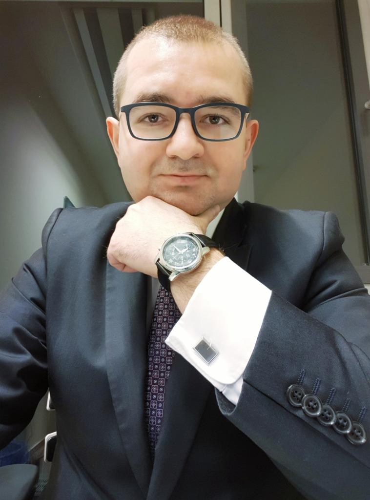 Portret mężczyzny w garniturze, z zegarkiem na ręku i spinkami do mankietów, opierającego brodę na dłoni, w tle jasna ściana.