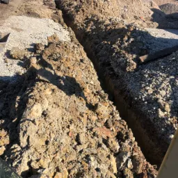 Wykopany wąski rów w ziemi, obok hałdy ziemi i gruzu, w tle domy jednorodzinne i błękitne niebo.