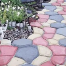 Ścieżka ogrodowa ułożona z nieregularnych, kolorowych betonowych płyt w odcieniach różu, szarości i beżu, prowadząca przez rabatę z białymi kwiatami i sadzonkami w doniczkach.