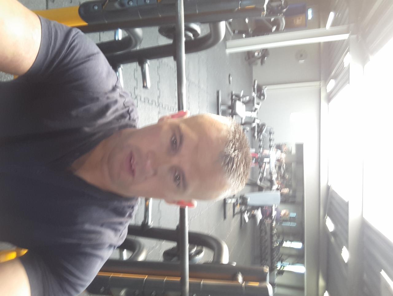 Mężczyzna w czarnej koszulce na tle wyposażenia siłowni, widoczne maszyny do ćwiczeń i podłoga z szarej gumy. Ujęcie z bliska, oświetlenie dzienne.