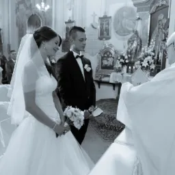 Czarno-białe zdjęcie z ceremonii ślubnej w kościele: panna młoda w białej sukni z bukietem, pan młody w czarnym smokingu, ksiądz w białej szacie udzielający błogosławieństwa, goście w tle.