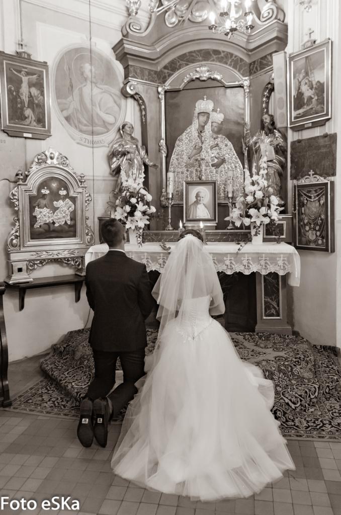 Czarno-białe zdjęcie pary młodej klęczącej przed ołtarzem w kościele, z widocznymi obrazami i figurami świętych oraz portretem Jana Pawła II.