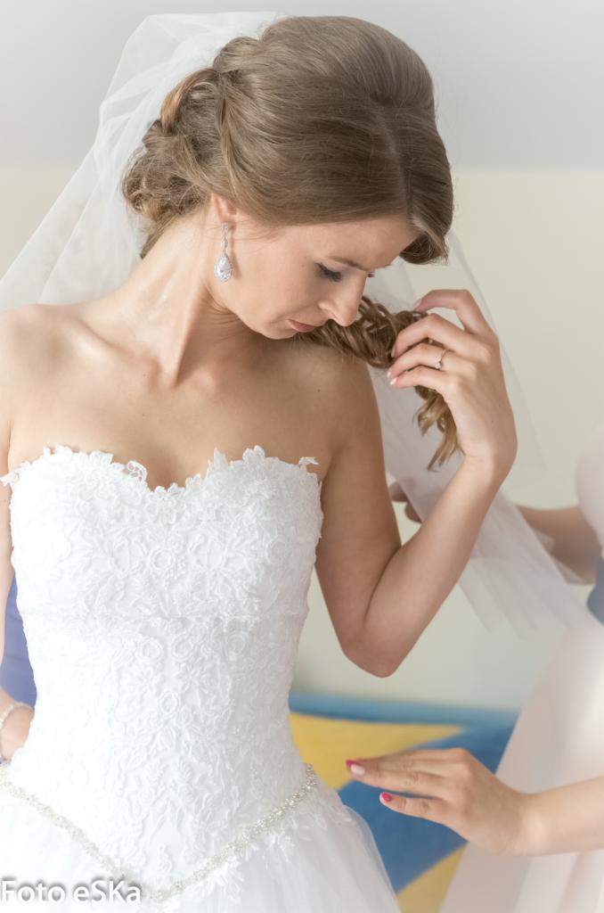 Panna młoda w koronkowej sukni bez ramiączek, z welonem i łezkowymi kolczykami, delikatnie dotyka loków, poprawiana przez kogoś podczas przygotowań ślubnych.