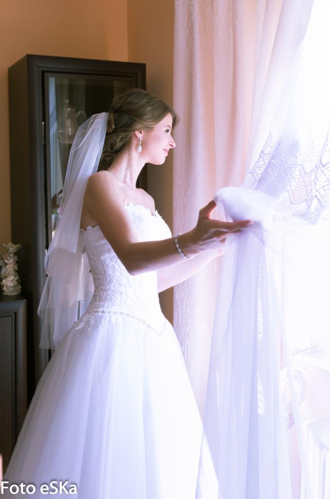 Panna młoda w białej sukni i welonie, z bransoletką na nadgarstku, spogląda przez okno, delikatnie odsuwając koronkową firankę.