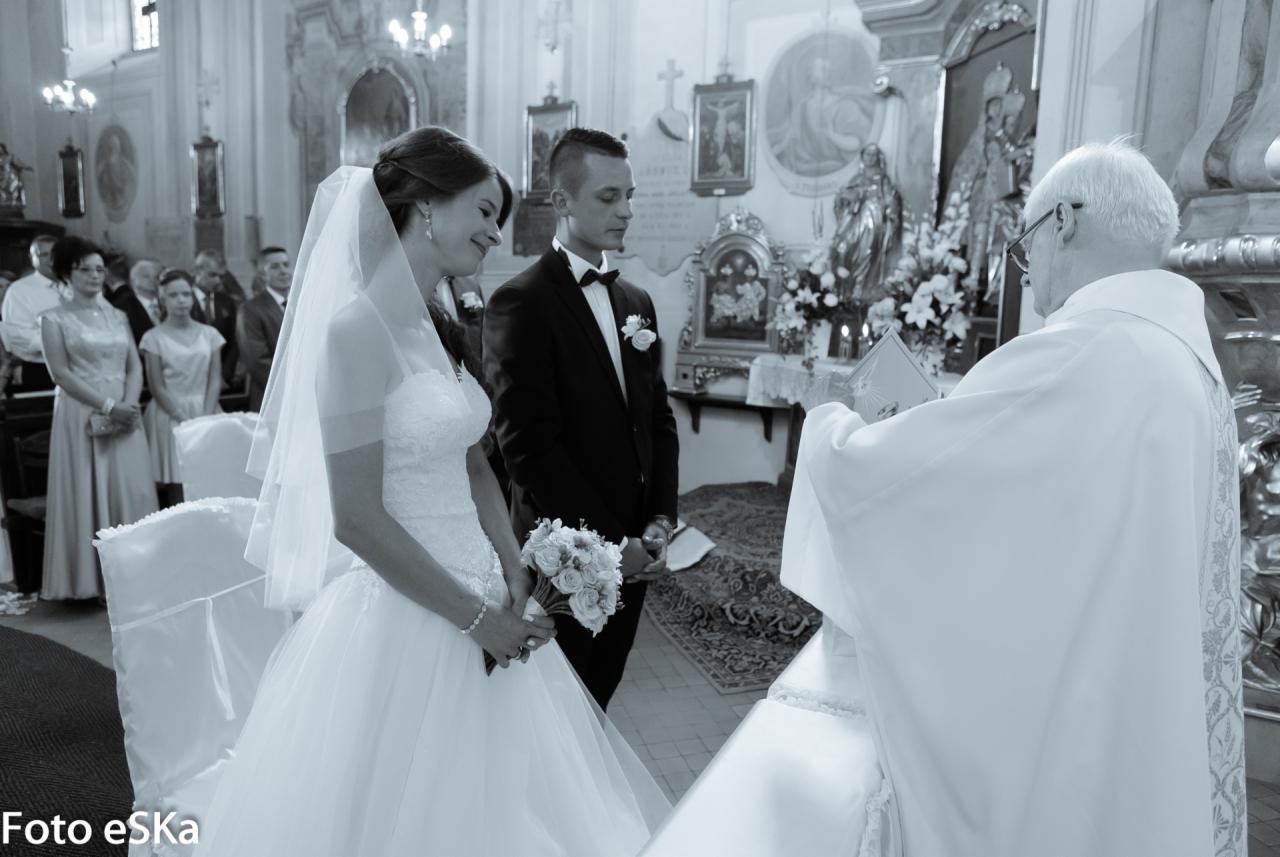 Czarno-białe zdjęcie z ceremonii ślubnej w kościele: panna młoda w białej sukni z bukietem, pan młody w czarnym smokingu, ksiądz w białej szacie udzielający błogosławieństwa, goście w tle.