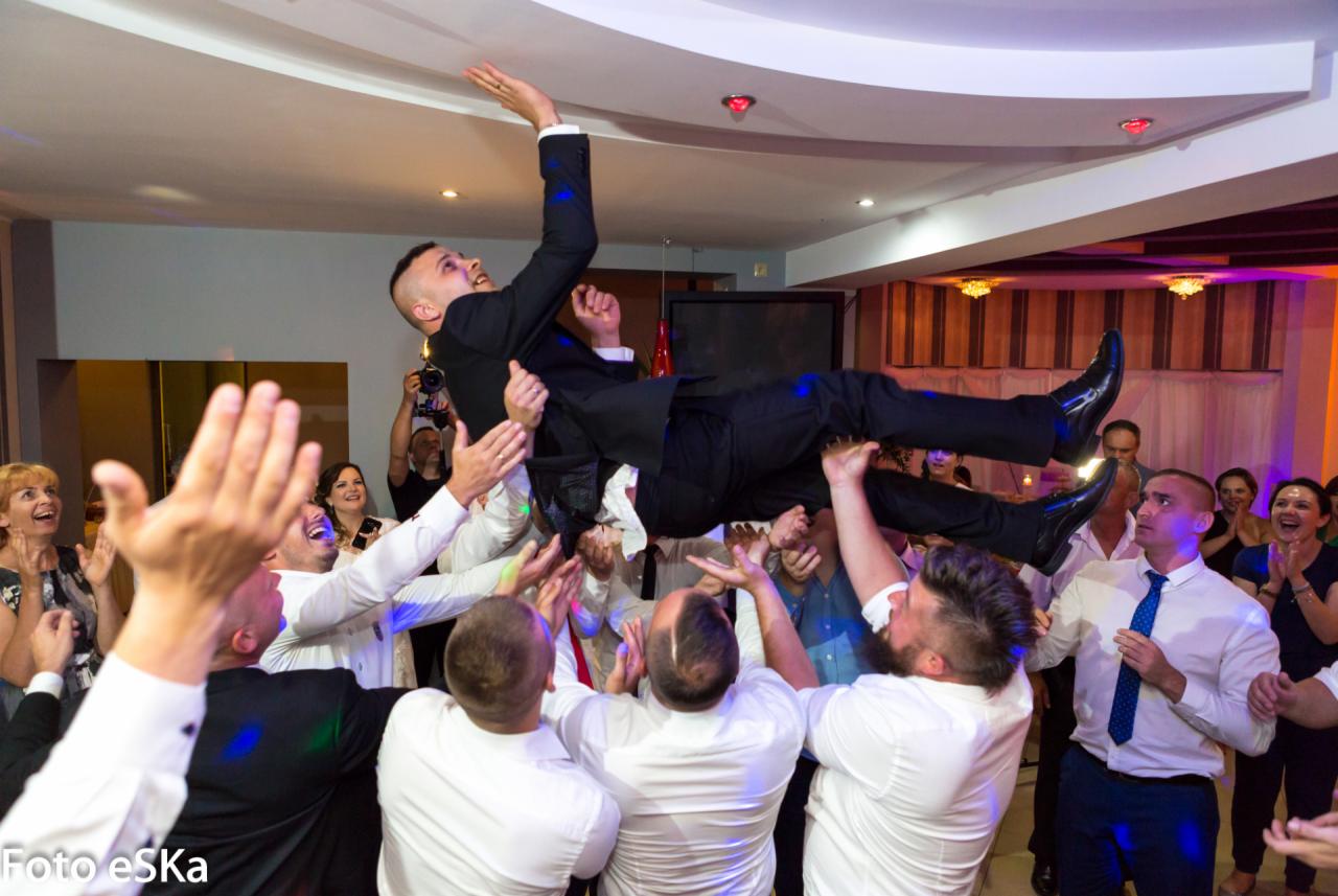 Uśmiechnięty pan młody w czarnym garniturze podrzucany przez gości weselnych podczas zabawy w sali z białym sufitem i dekoracyjnym oświetleniem, w tle fotograf robiący zdjęcia.