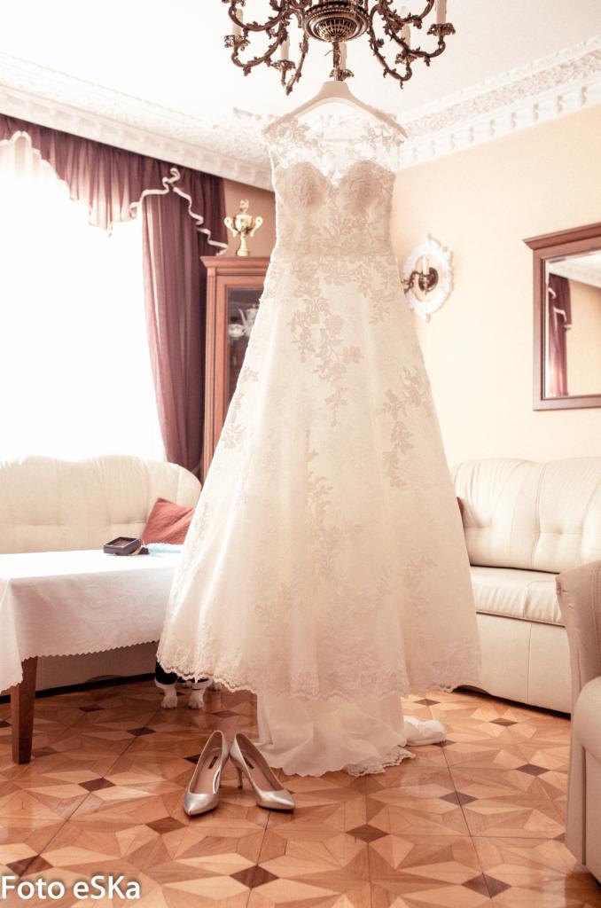 Delikatna suknia ślubna z koronkowymi aplikacjami, wisząca na wieszaku pod ozdobnym żyrandolem, z srebrnymi szpilkami u dołu i fragmentem wnętrza w tle.