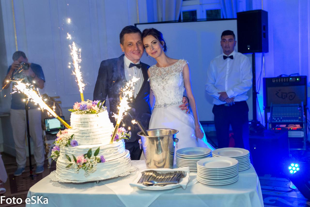 Para młoda pozuje za tortem weselnym z zimnymi ogniami, w tle fotograf i obsługa, oświetlenie w odcieniach błękitu.