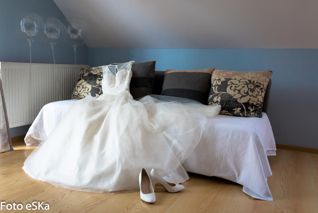 Biała suknia ślubna z tiulowym dołem i koronkową górą, leżąca na białej sofie z dekoracyjnymi poduszkami, obok białe buty na wysokim obcasie, w tle niebieska ściana i balony.