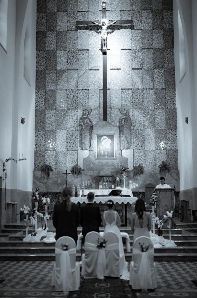 Czarno-białe zdjęcie ślubu w kościele z mozaikową ścianą za ołtarzem, gdzie para młoda stoi tyłem do widza, a ksiądz prowadzi ceremonię.