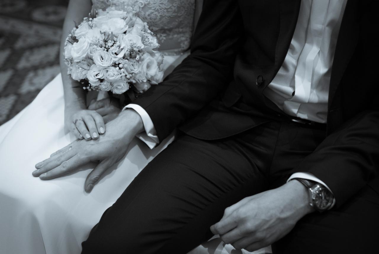 Czarno-białe zbliżenie dłoni panny młodej z bukietem i pana młodego w garniturze, z widocznym zegarkiem na jego nadgarstku.