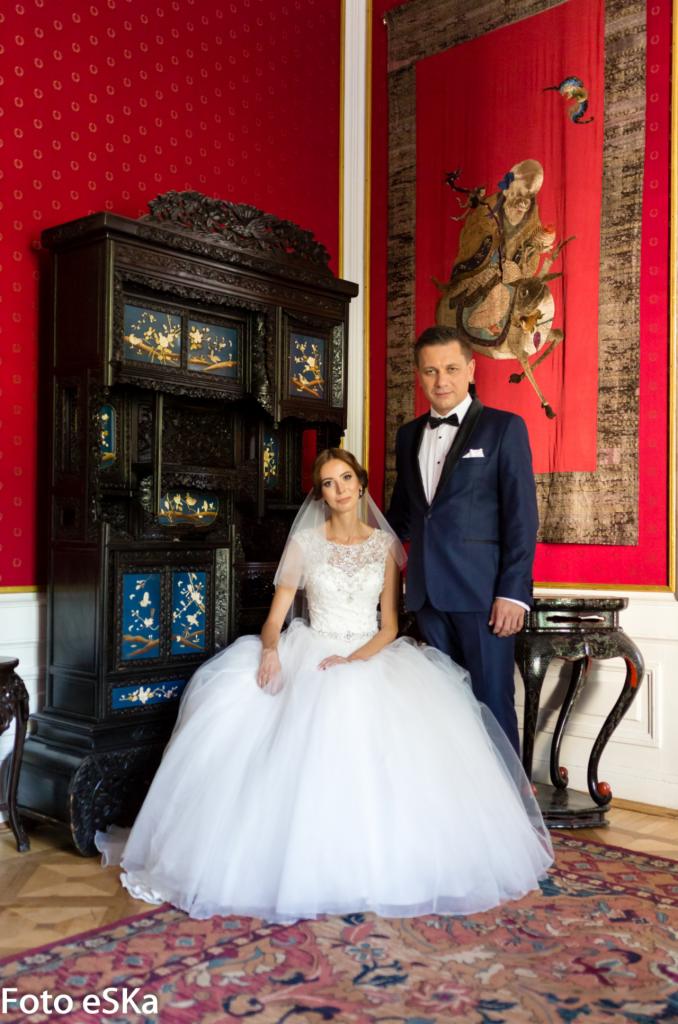 Para młoda: panna młoda w białej sukni siedzi, pan młody w granatowym garniturze stoi, na tle czerwonej ściany z orientalnym gobelinem i ciemnego, zdobionego kredensu.