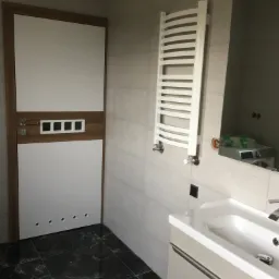 Wykończona łazienka z czarną marmurową podłogą, białą umywalką z szafką, grzejnikiem drabinkowym i drzwiami z drewnianymi akcentami.