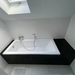 Biała wanna obudowana czarnymi płytkami w nowoczesnej łazience z oknem dachowym, widoczne białe płytki ścienne z delikatną fakturą i armatura.