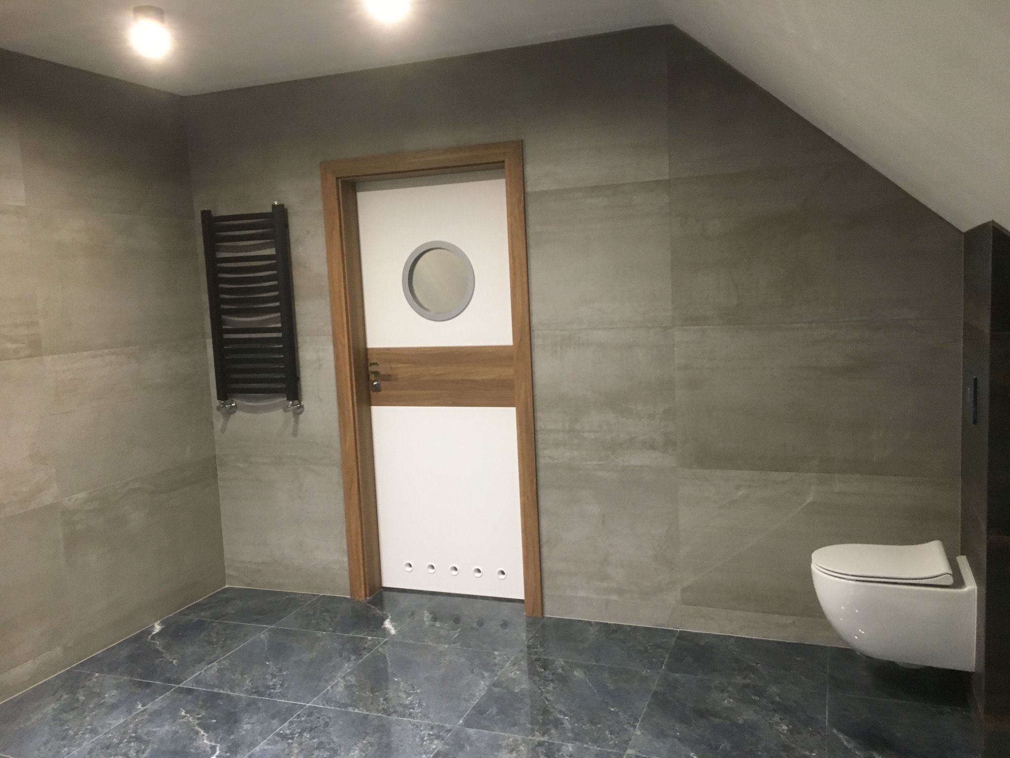 Nowoczesna łazienka na poddaszu z szarymi płytkami, grzejnikiem drabinkowym, podwieszaną toaletą i białą, nietypową drzwi z okrągłym okienkiem.