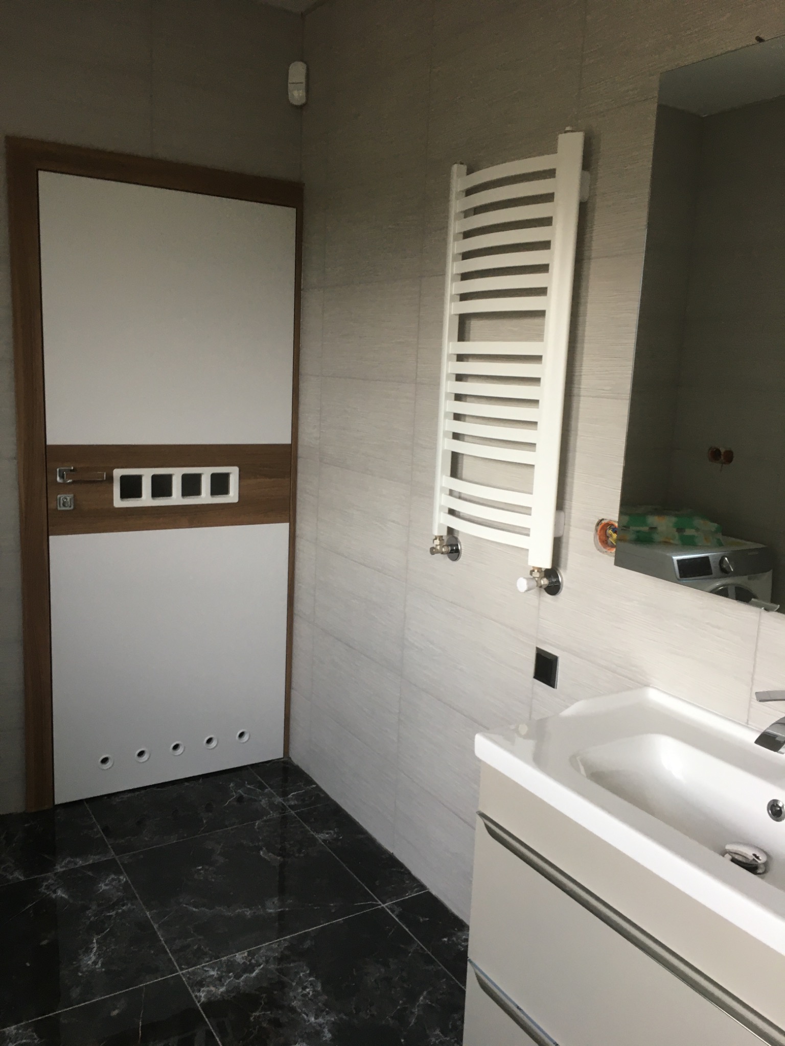 Wykończona łazienka z czarną marmurową podłogą, białą umywalką z szafką, grzejnikiem drabinkowym i drzwiami z drewnianymi akcentami.