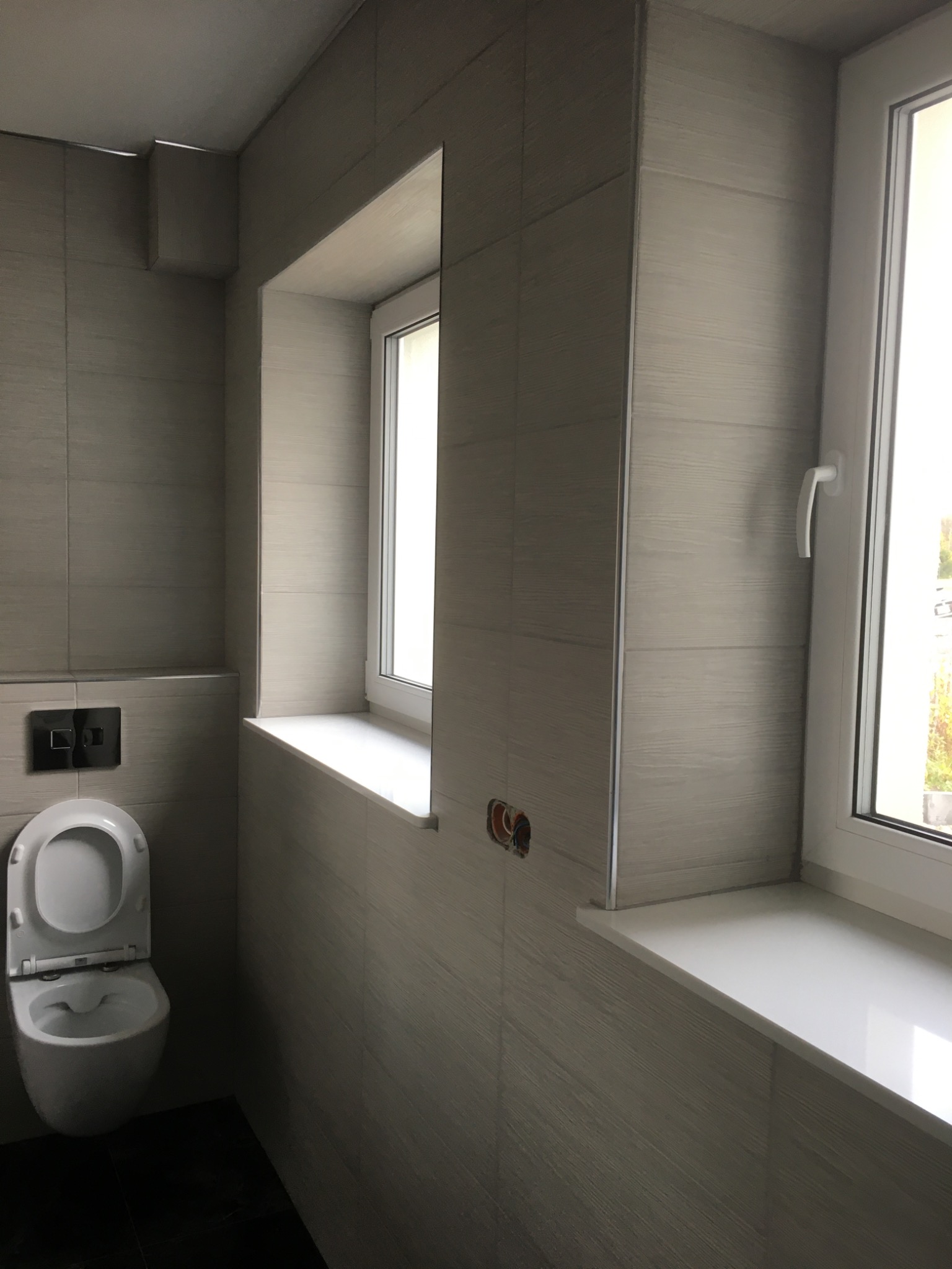 Nowoczesna łazienka z podwieszaną toaletą, płytkami imitującymi drewno i oknami z białymi parapetami. Widoczne wystające kable elektryczne. Ciemna podłoga.