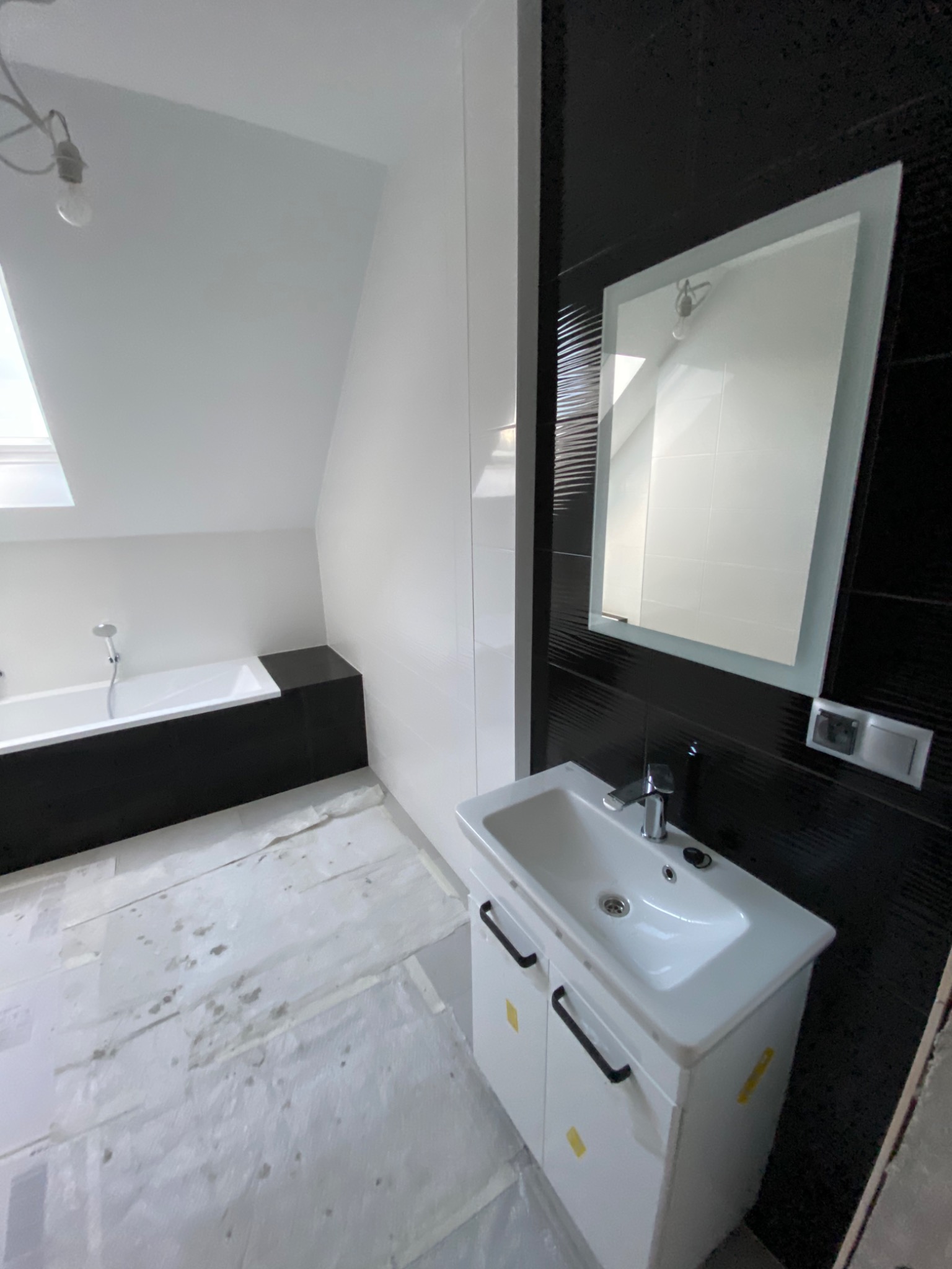 Nowoczesna łazienka na poddaszu w trakcie prac wykończeniowych: biała wanna z czarną obudową, umywalka z szafką, czarne płytki na ścianie i lustro. Podłoga zabezpieczona folią malarską.