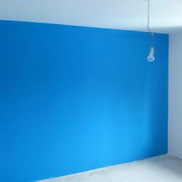 Marcin-Gładzie -Malowanie - Świeżo pomalowana na niebiesko ściana w pokoju, z widoczną żarówką zwisającą z sufitu na kablu, surowy betonowy podkład pod podłogę.