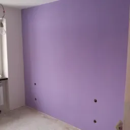 Świeżo pomalowana ściana w odcieniach fioletu z widocznymi otworami po gniazdkach elektrycznych, surowy beton na podłodze, żarówka zwisająca z sufitu.