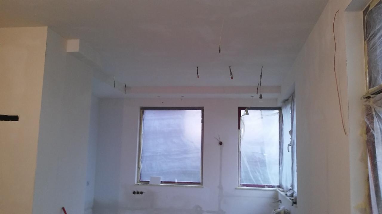 Wnętrze w trakcie remontu: białe ściany, przygotowane do malowania, okna zabezpieczone folią malarską, widoczne wystające przewody elektryczne.