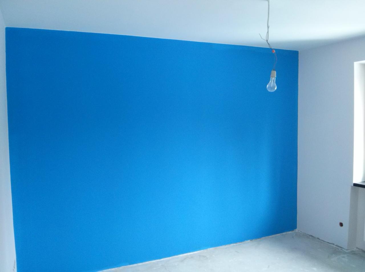 Świeżo pomalowana na niebiesko ściana w pokoju, z widoczną żarówką zwisającą z sufitu na kablu, surowy betonowy podkład pod podłogę.