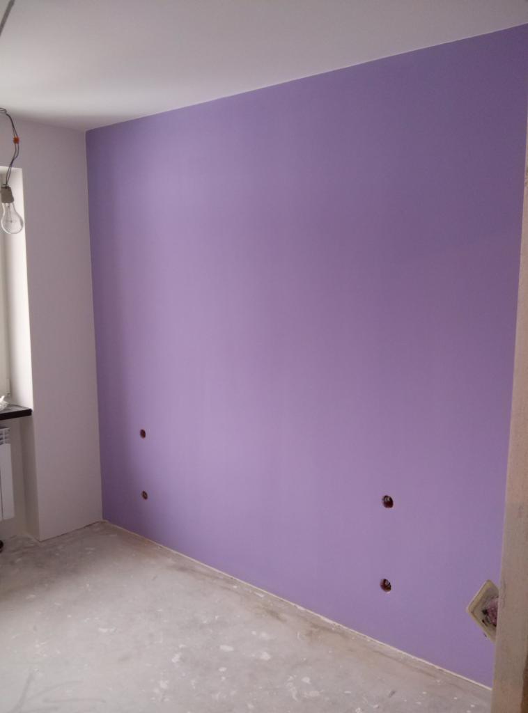 Świeżo pomalowana ściana w odcieniach fioletu z widocznymi otworami po gniazdkach elektrycznych, surowy beton na podłodze, żarówka zwisająca z sufitu.