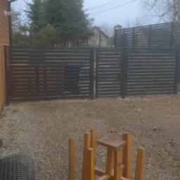 Nowoczesne, poziome ogrodzenie metalowe w kolorze grafitowym z furtką, widoczne z perspektywy podwórza z żwirowym podłożem, z widoczną konstrukcją drewnianą prawdopodobnie używaną podczas montażu...