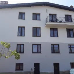 Trzykondygnacyjny budynek po remoncie z jasnoszarą elewacją, ciemnymi oknami i balkonem z metalową balustradą, widoczne płyty styropianowe u podstawy.