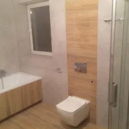 Nowoczesna łazienka z wanną obudowaną drewnem, wiszącą toaletą i kabiną prysznicową z chromowanymi detalami, wykończona jasnymi płytkami imitującymi drewno i kamień.