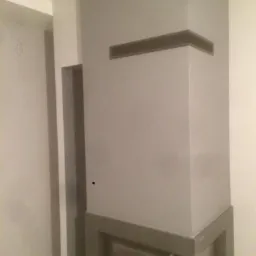 Szary, narożny kominek z wkładem, obudowany płytami w odcieniach szarości, z poziomym otworem wentylacyjnym w górnej części i prostokątną wnęką z boku.