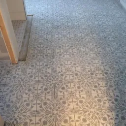 Ułożone szaro-białe płytki podłogowe z dekoracyjnym wzorem w kształcie rozety, widoczne krzyżyki dystansowe między płytkami, fragment niewykończonej ściany i progu.