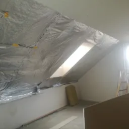 Poddasze w trakcie prac izolacyjnych, widoczna folia aluminiowa z taśmą mocującą, fragment okna dachowego i drabina oparta o ścianę.