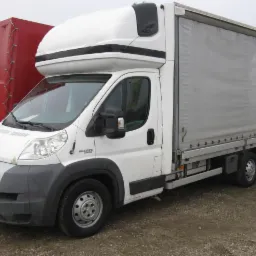 Biały samochód dostawczy Fiat Ducato z wysoką kabiną i szarą plandeką na tle przyczepy z czerwoną plandeką oraz reklamy Agro-Mar.