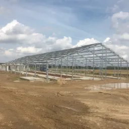 Stalowa konstrukcja hali w trakcie budowy, widoczne fundamenty i niebo z chmurami w tle.