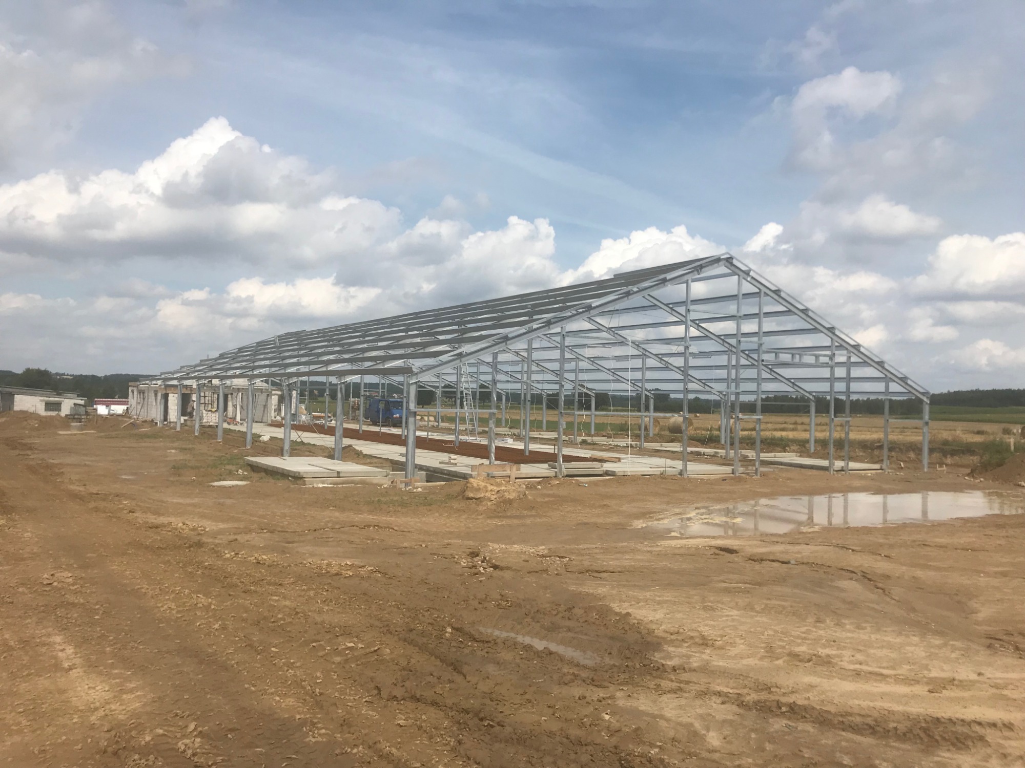 Stalowa konstrukcja hali w trakcie budowy, widoczne fundamenty i niebo z chmurami w tle.